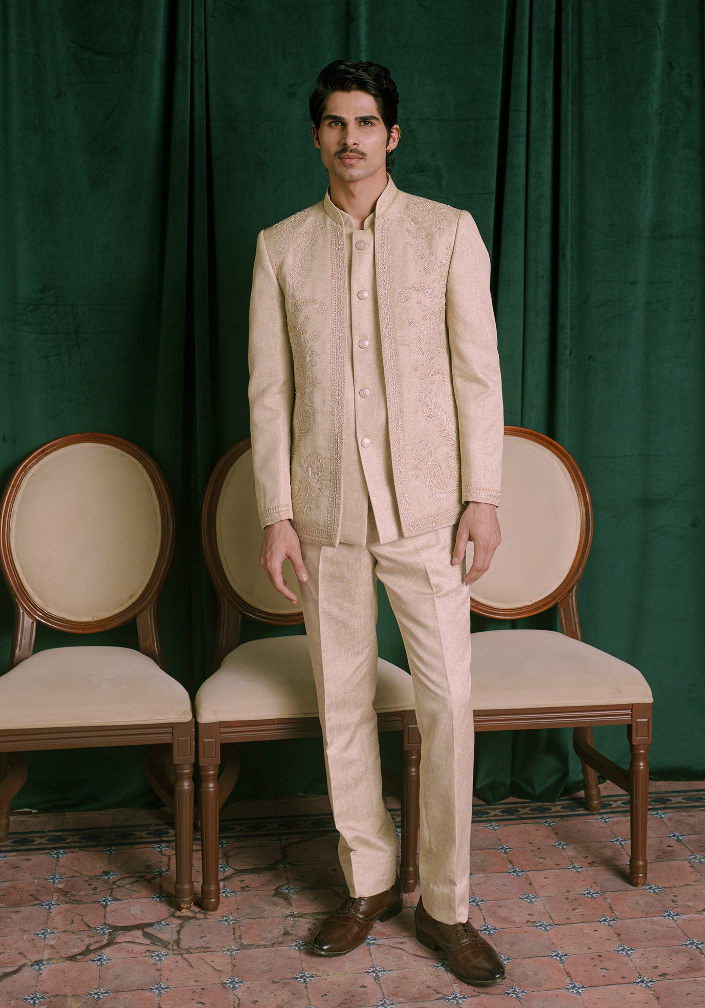 A man wearing a beige colored embroidered Indian bandhgala jacket and matching trousers, standing in front of a dark green curtain with two wooden chairs behind him.