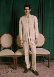 A man wearing a beige colored embroidered Indian bandhgala jacket and matching trousers, standing in front of a dark green curtain with two wooden chairs behind him.