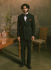 A man wearing a black double-breasted tuxedo with satin peak lapels, a black bow tie, a white shirt, and a red pocket square, standing in a dimly lit, vintage-style room with a table, candles, and a chair.