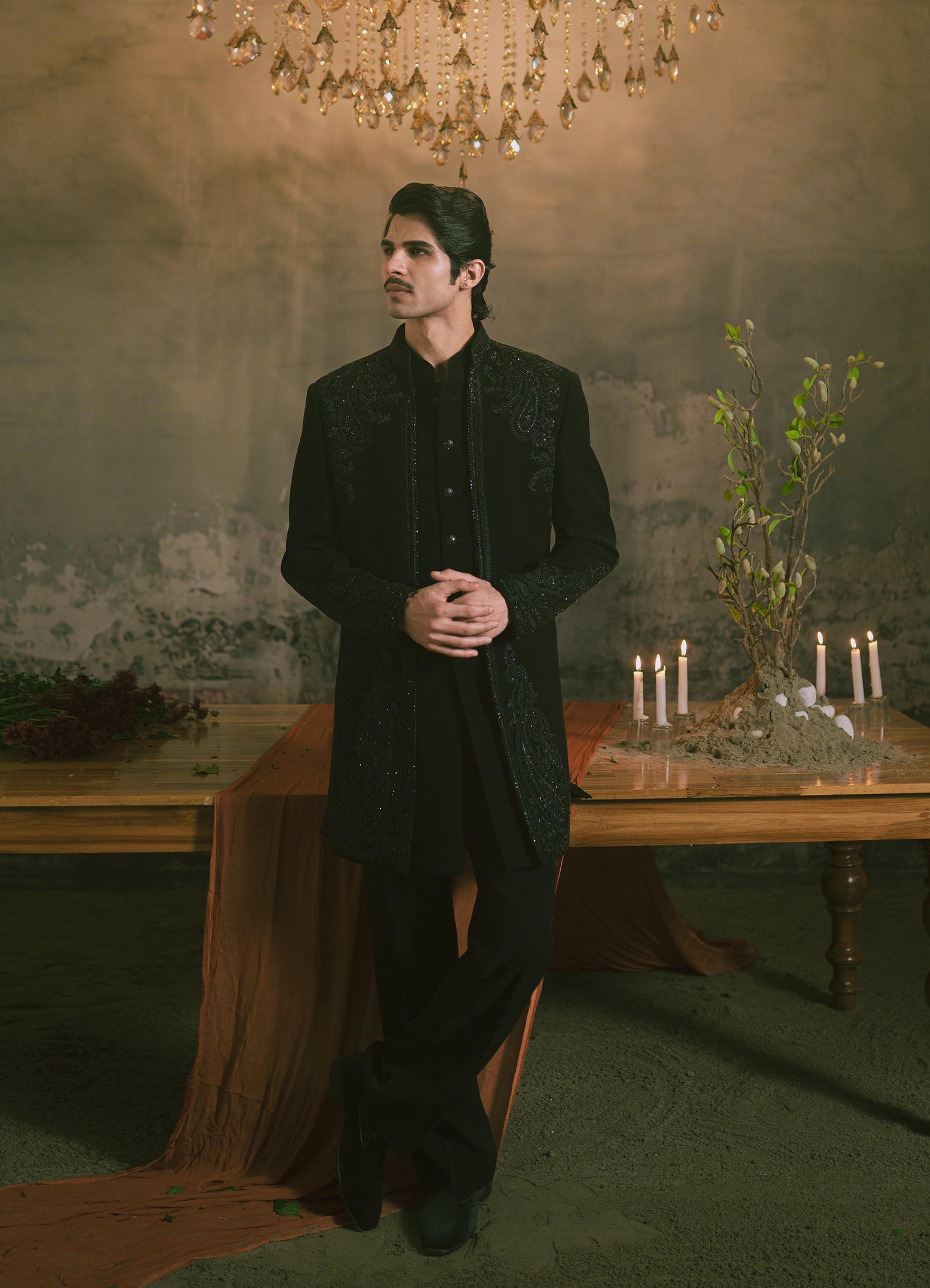 A man wearing a black embroidered traditional formal jacket and trousers stands in a dimly lit room with a chandelier, rustic wall, and a table with lit candles and dried flowers.