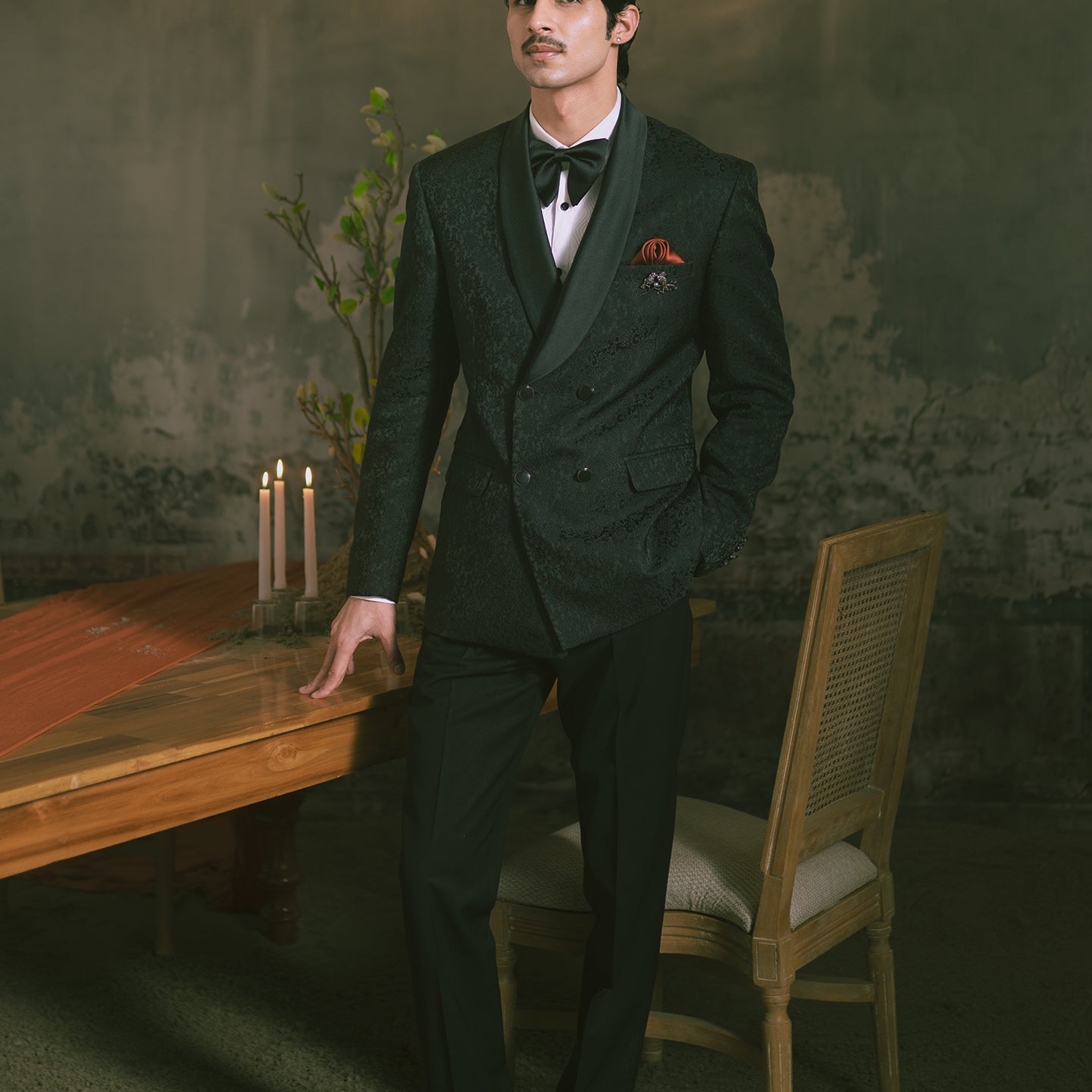 A man wearing a dark double-breasted suit with a subtle pattern, a white shirt, a black bow tie, and a red pocket square.