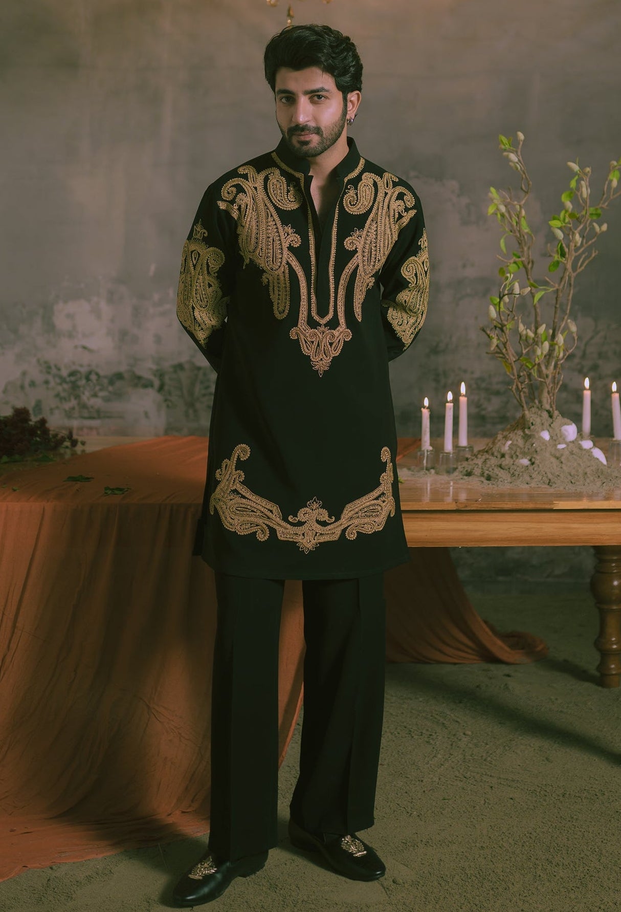 A man wearing a black traditional kurta with ornate gold embroidery and matching black trousers, standing in a moody indoor setting with a chandelier, draped fabric, and candles on a table.