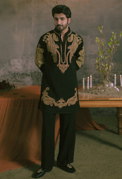 A man wearing a black traditional kurta with ornate gold embroidery and matching black trousers, standing in a moody indoor setting with a chandelier, draped fabric, and candles on a table.