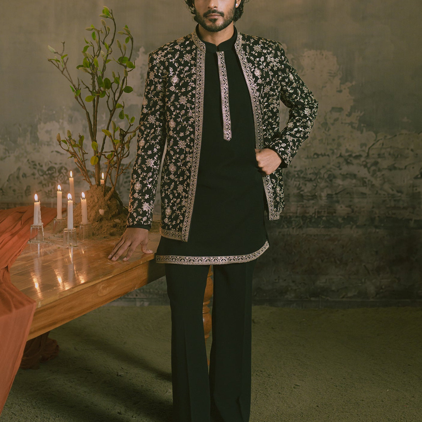 A man wearing a black traditional outfit with ornate golden embroidery and a black patterned jacket, standing in a rustic room with a chandelier, table with candles, and a plant.