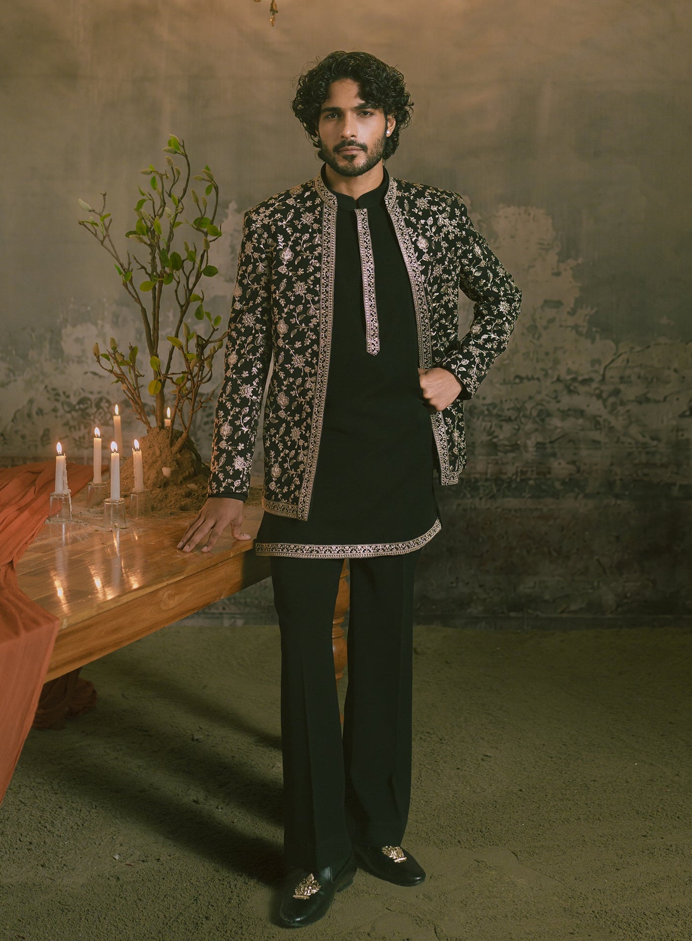 A man wearing a black traditional outfit with ornate golden embroidery and a black patterned jacket, standing in a rustic room with a chandelier, table with candles, and a plant.