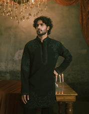 A man wearing a black long-sleeve short kurta with intricate embroidery & wide bottom trousers.