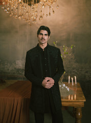 A man wearing a black embroidered sherwani jacket over a black kurta and trousers, standing in a dimly lit, elegant room with a chandelier, a wooden table with lit candles, and a draped brown cloth.