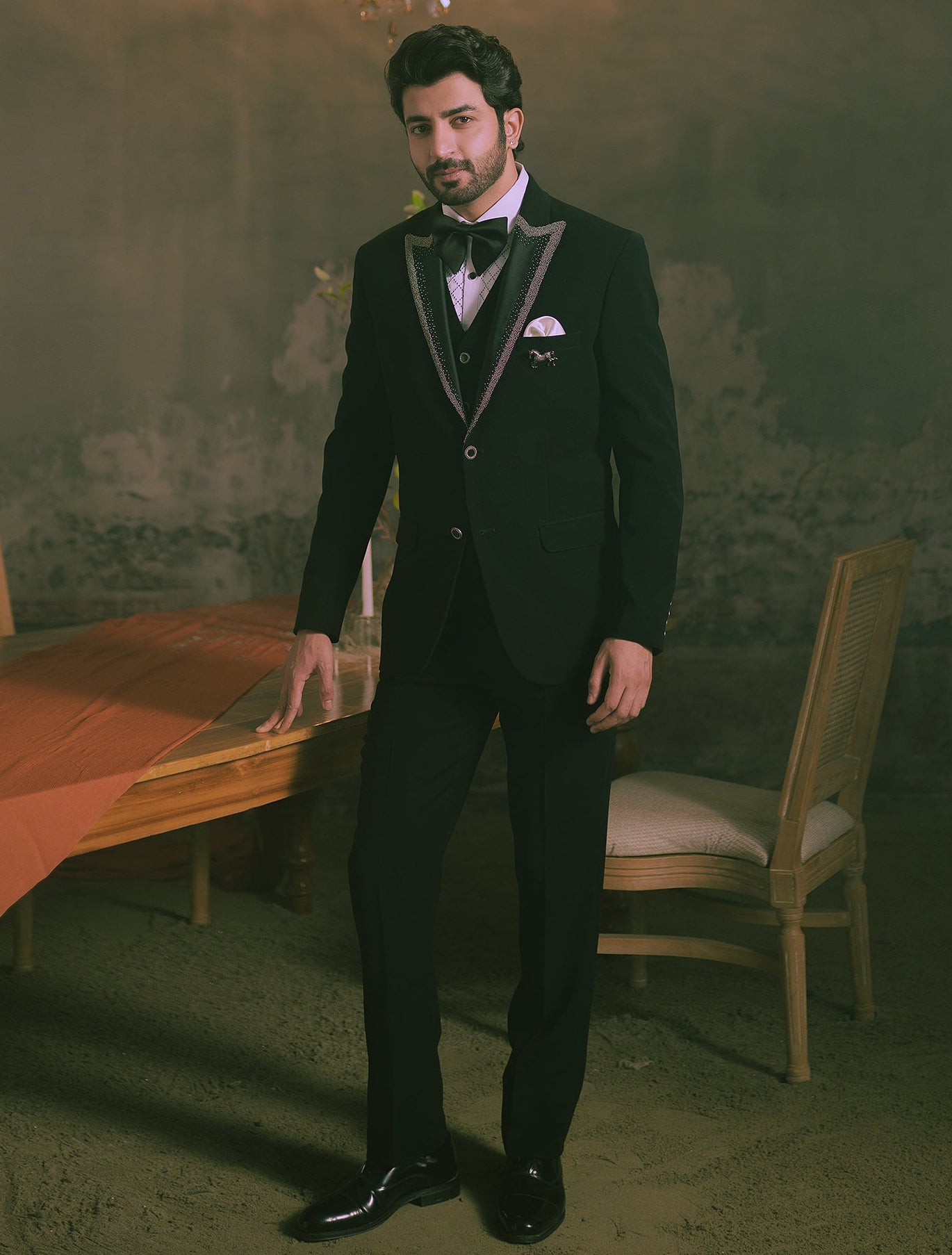A man wearing a black tuxedo with silver peak lapels, a white shirt, a black bow tie, and a white pocket square stands in a dimly lit, vintage-style room with a chandelier and a table draped in orange fabric.