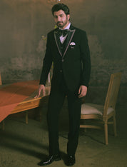 A man wearing a black tuxedo with silver peak lapels, a white shirt, a black bow tie, and a white pocket square stands in a dimly lit, vintage-style room with a chandelier and a table draped in orange fabric.