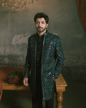 A man is wearing a dark green heavily embellished sherwani jacket with sequins and embroidery.