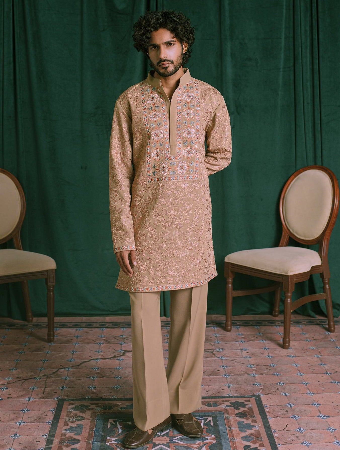 A man wearing a brown embroidered kurta and matching trousers stands in front of a dark green curtain backdrop, with two wooden chairs on either side and a patterned rug beneath his feet.