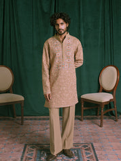 A man wearing a brown embroidered kurta and matching trousers stands in front of a dark green curtain backdrop, with two wooden chairs on either side and a patterned rug beneath his feet.