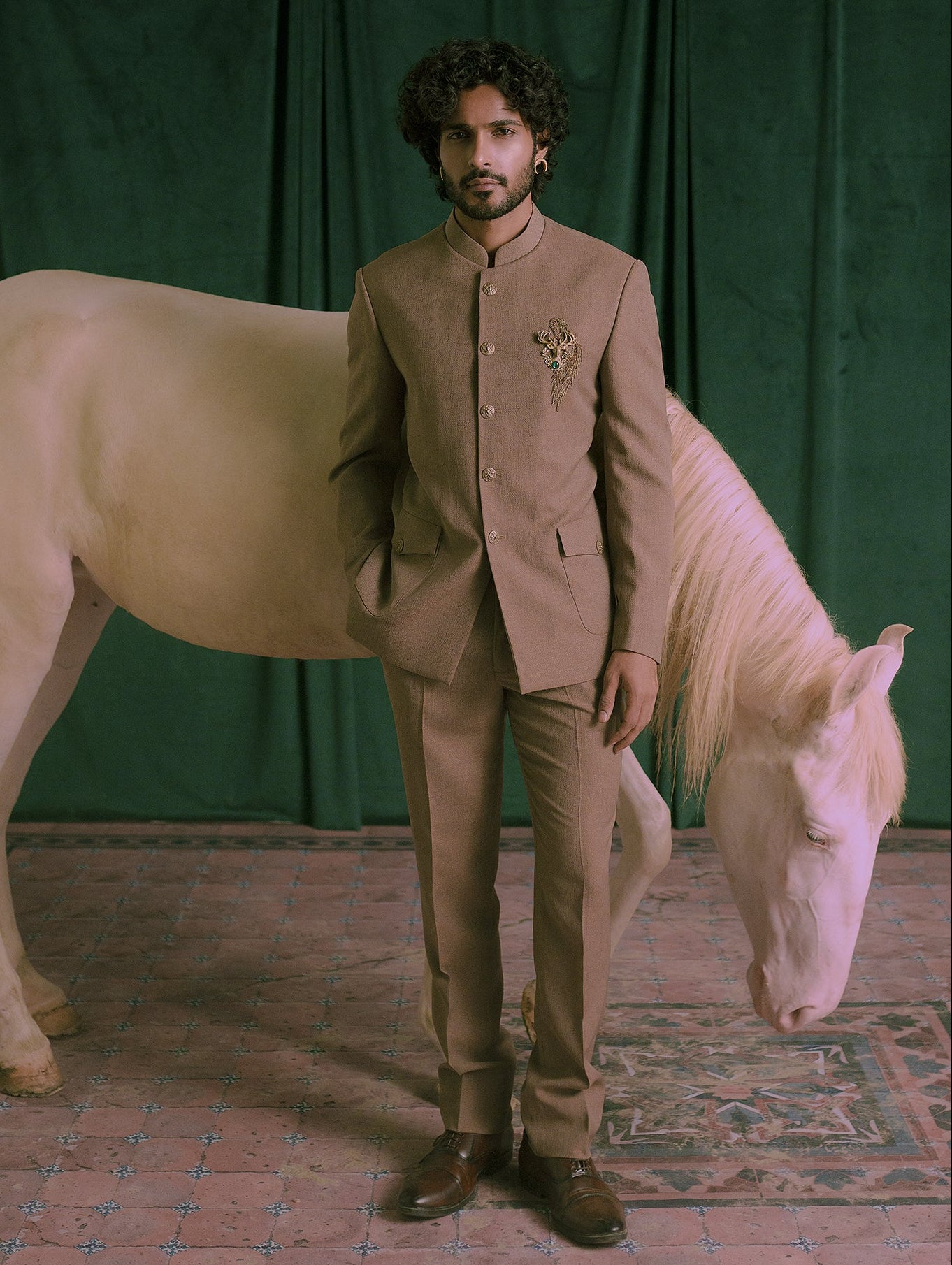 A man wearing a brown traditional South Asian suit stands next to a white horse in front of a dark green curtain backdrop.