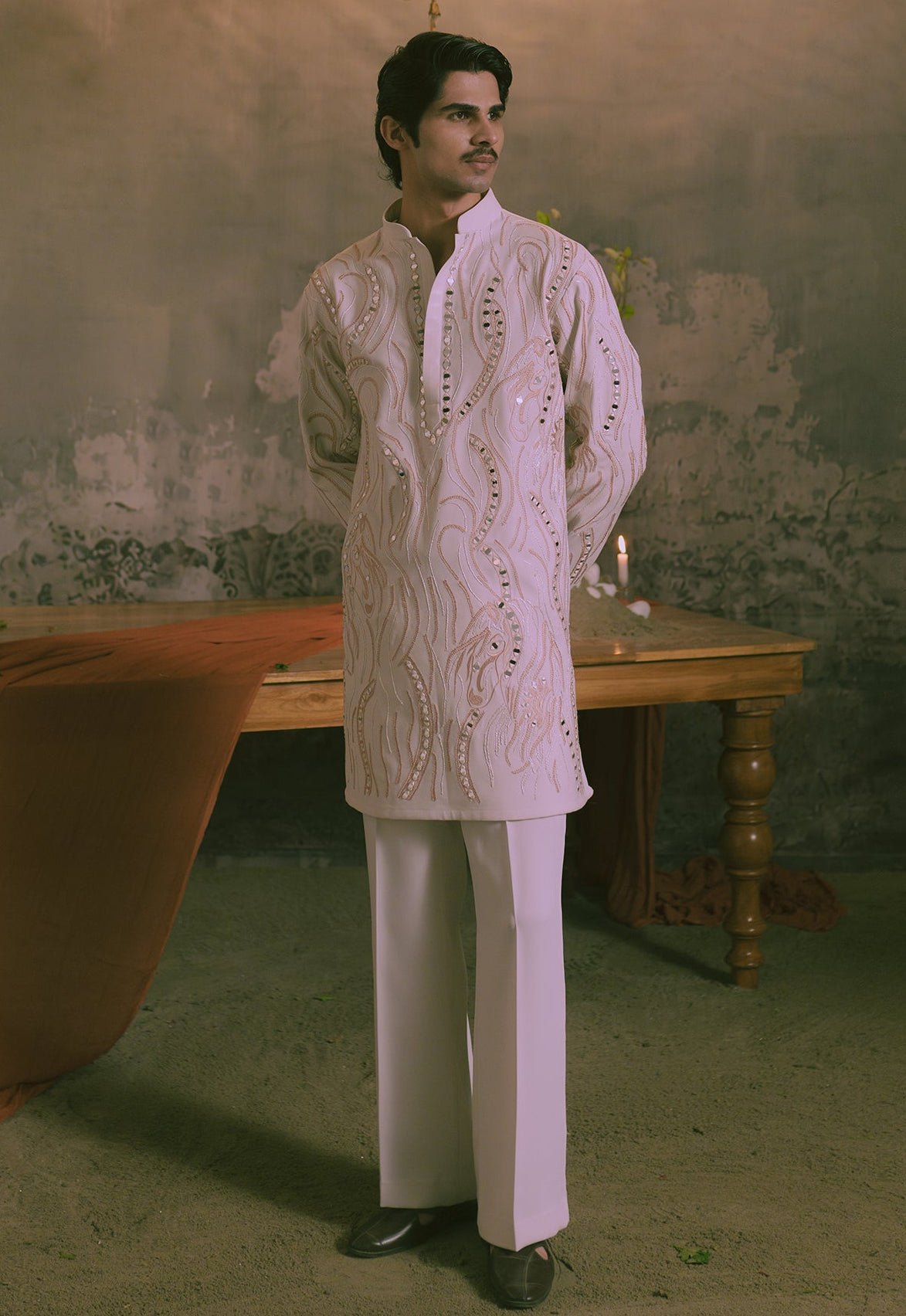 A man wearing a cream embroidered kurta and white trousers stands in a rustic room with a textured wall, a wooden table, and a draped orange fabric.