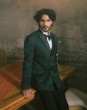 A man with curly dark hair and a beard is wearing a dark green textured tuxedo jacket with black satin lapels, a black bow tie, and a white shirt.