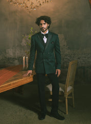 A man is wearing a dark green double-breasted tuxedo jacket with black satin lapels, a black bow tie, and black trousers.