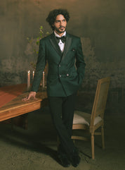 A man is wearing a dark green double-breasted tuxedo with black satin lapels, a black bow tie, and black trousers.