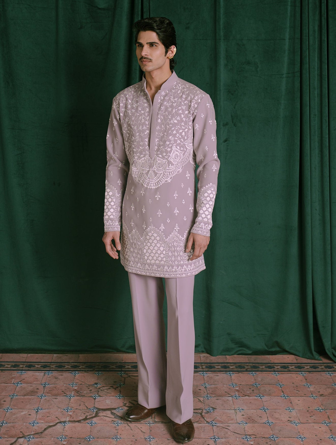 A man modeling a light lavender traditional Indian outfit consisting of a long-sleeve embroidered kurta and matching trousers, standing against a dark green velvet curtain backdrop.