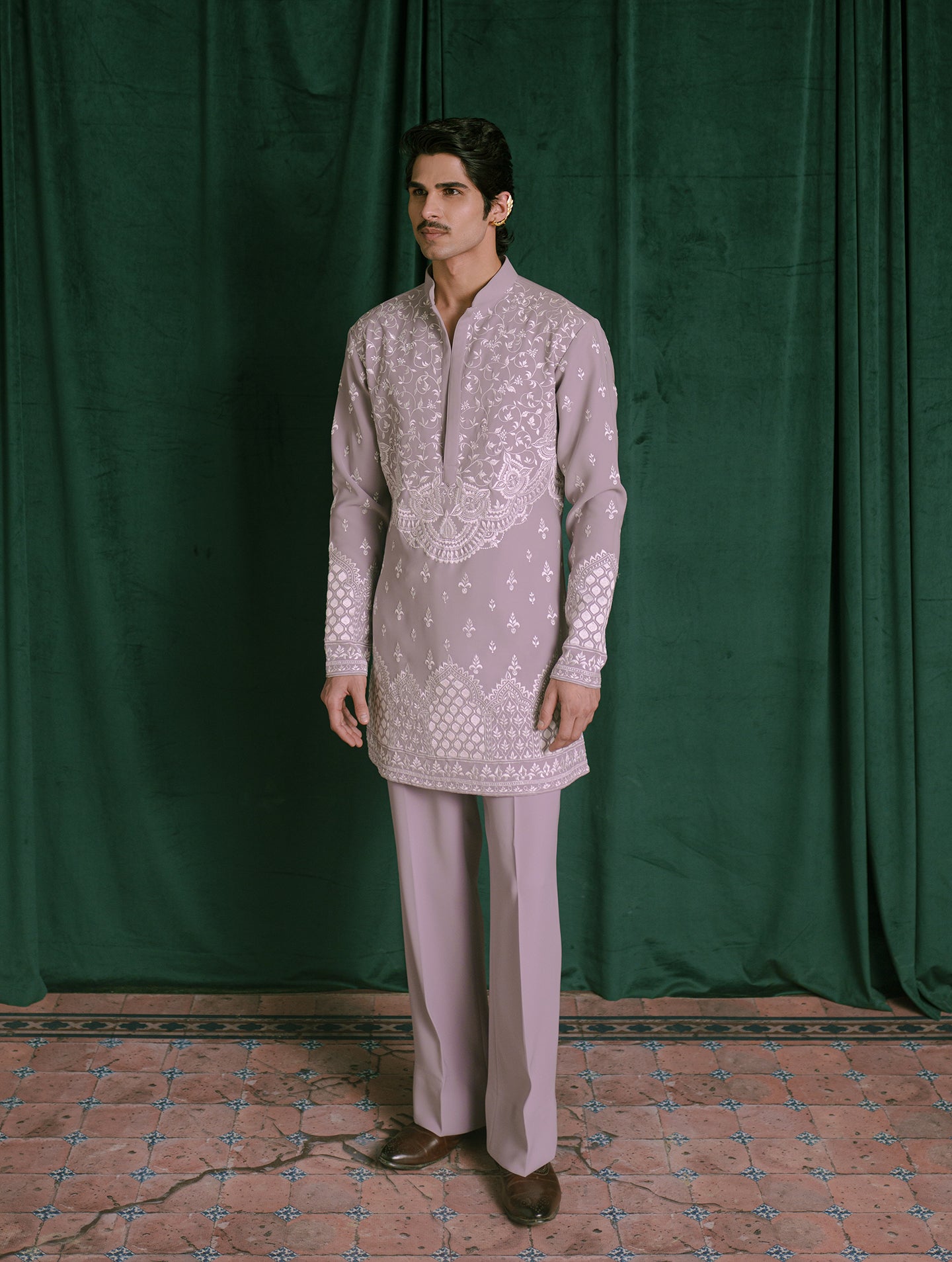 A man modeling a light lavender traditional Indian outfit consisting of a long-sleeve embroidered kurta and matching trousers, standing against a dark green velvet curtain backdrop.