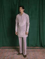 A man modeling a light lavender traditional Indian outfit consisting of a long-sleeve embroidered kurta and matching trousers, standing against a dark green velvet curtain backdrop.