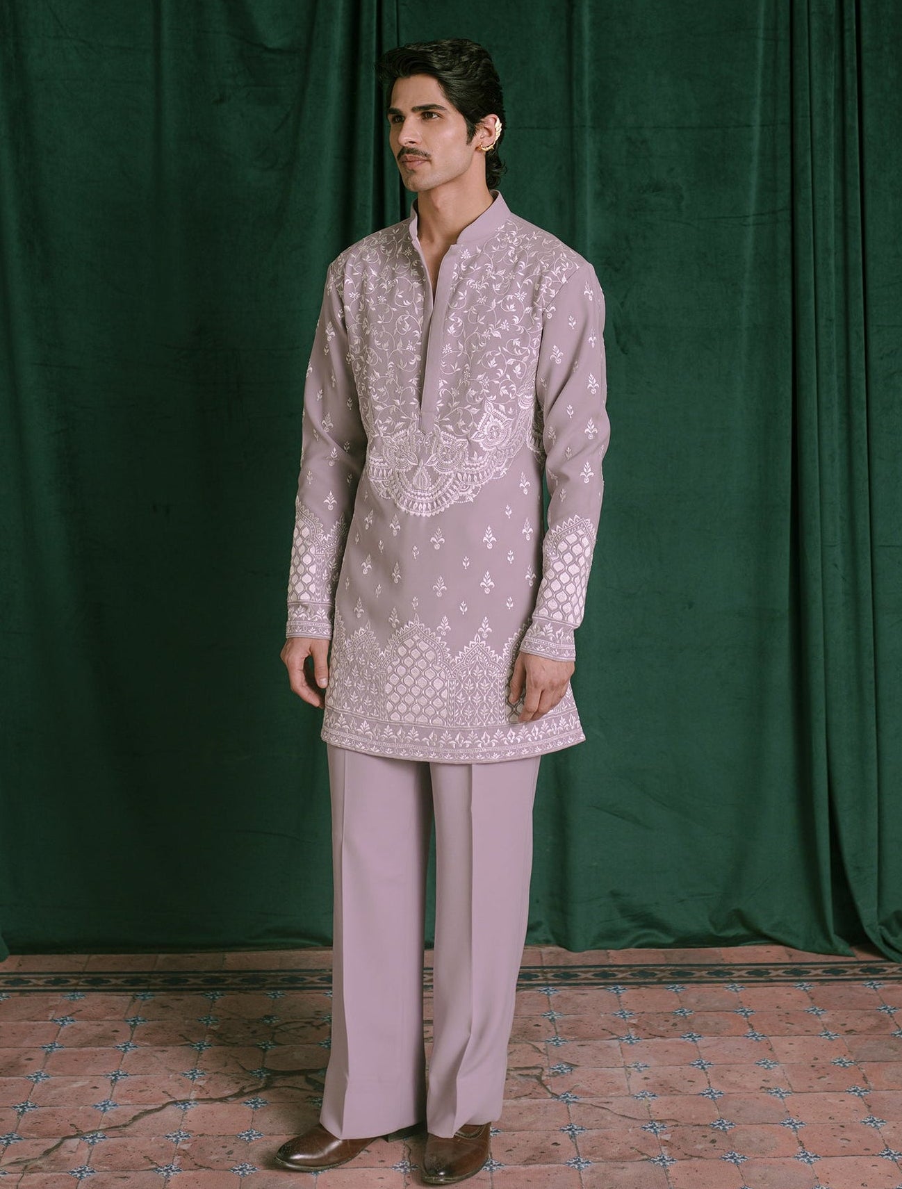 A man modeling a light lavender traditional Indian outfit consisting of a long-sleeve kurta with white embroidery and matching trousers, standing in front of a dark green velvet curtain.