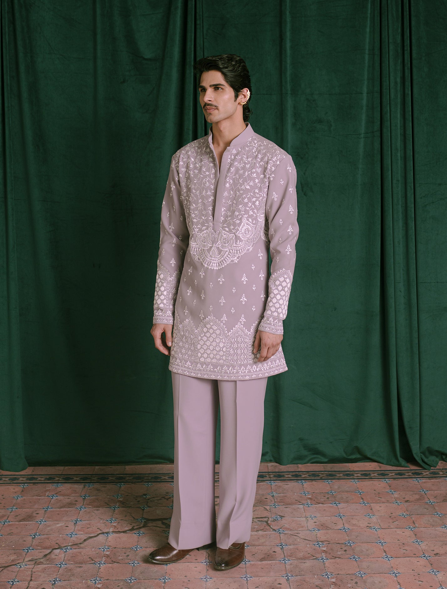 A man modeling a light lavender traditional Indian outfit consisting of a long-sleeve kurta with white embroidery and matching trousers, standing in front of a dark green velvet curtain.