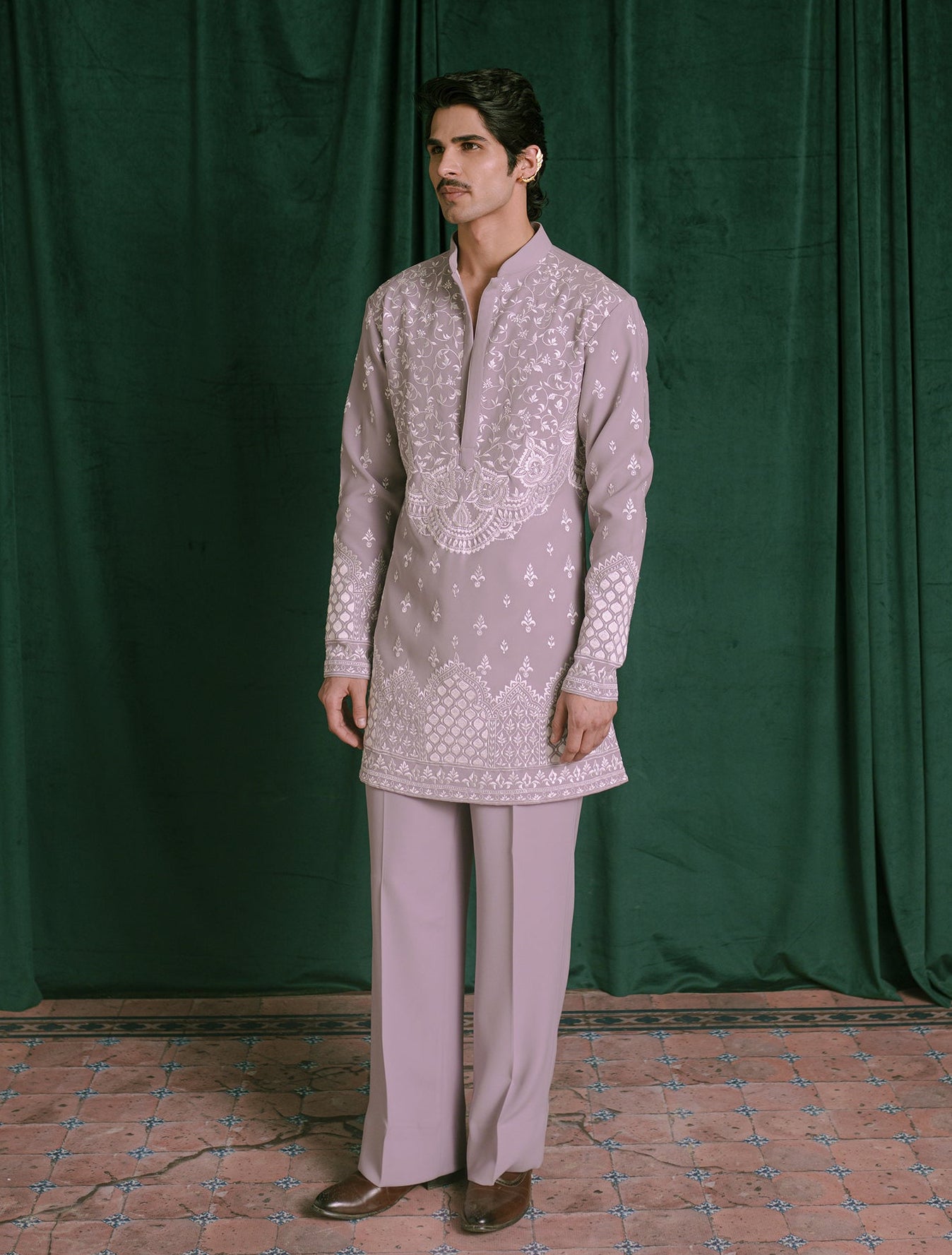 A man modeling a light lavender traditional Indian outfit consisting of a long-sleeve kurta with white embroidery and matching trousers, standing in front of a dark green velvet curtain.