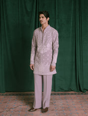 A man modeling a light lavender traditional Indian outfit consisting of a long-sleeve kurta with white embroidery and matching trousers, standing in front of a dark green velvet curtain.