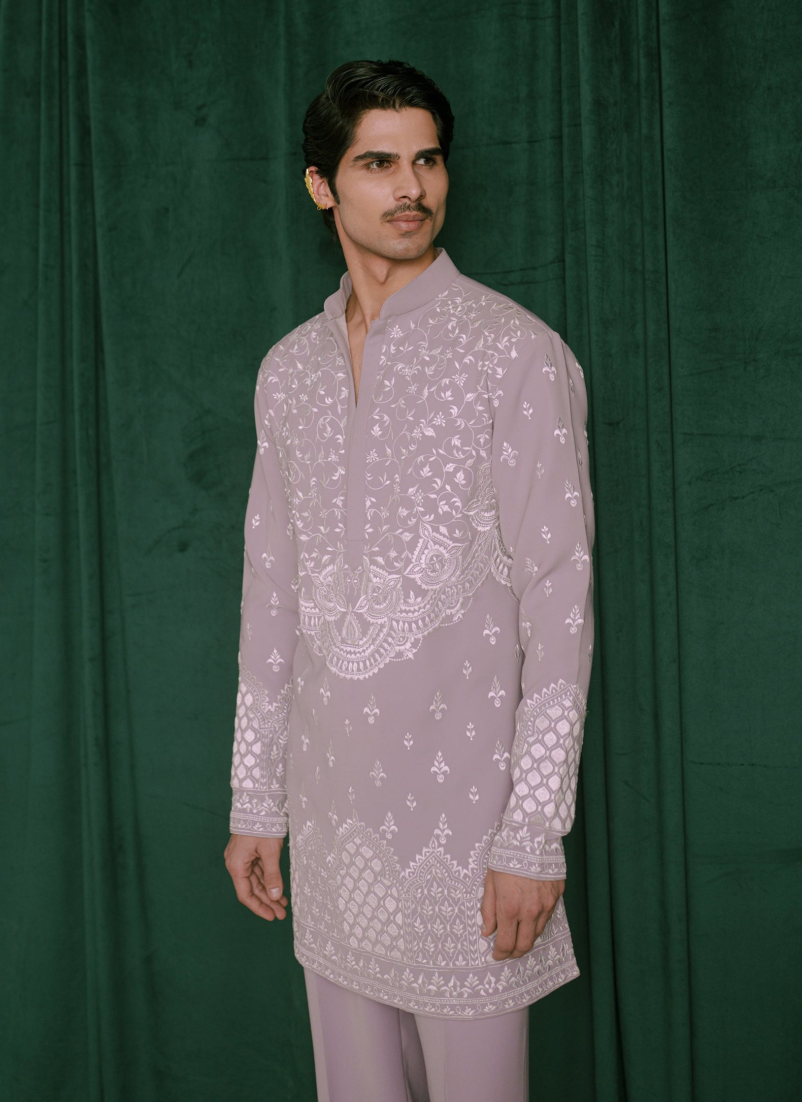 A man modeling a light lavender embroidered kurta with white floral and geometric patterns, paired with matching trousers, standing against a dark green velvet backdrop.