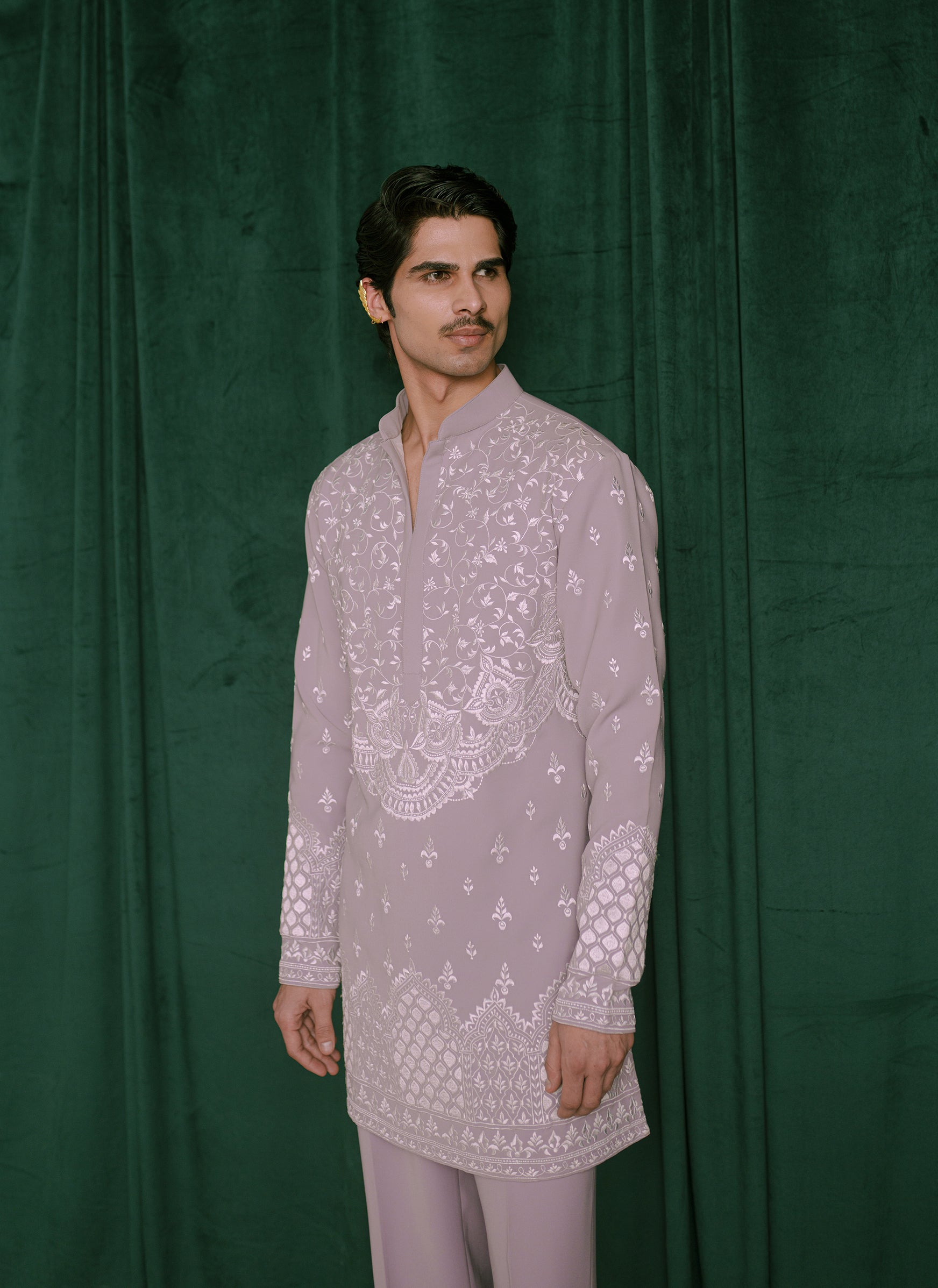 A man modeling a light lavender embroidered kurta with white floral and geometric patterns, paired with matching trousers, standing against a dark green velvet backdrop.