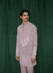 A man modeling a light lavender embroidered kurta with white floral and geometric patterns, paired with matching trousers, standing against a dark green velvet backdrop.