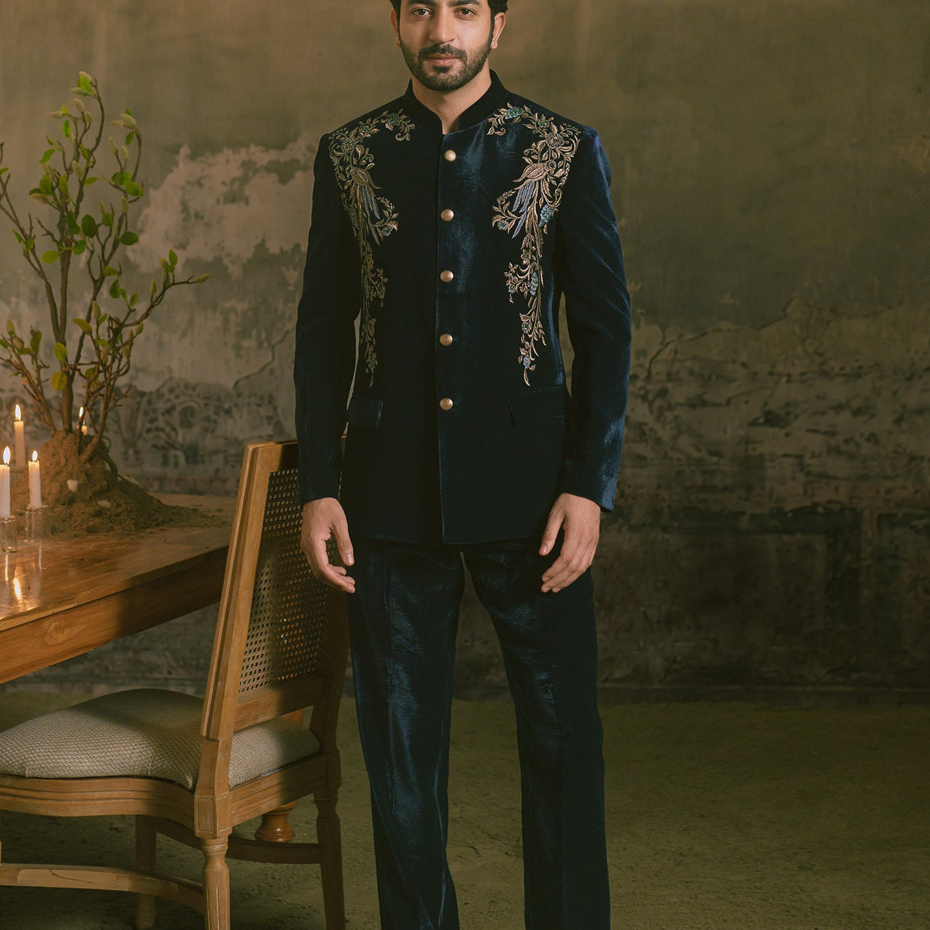 A man wearing a dark navy blue embroidered traditional Indian formal suit with gold buttons and silver mirror-work details on the chest, standing in a dimly lit room with a chandelier, table, and candles.