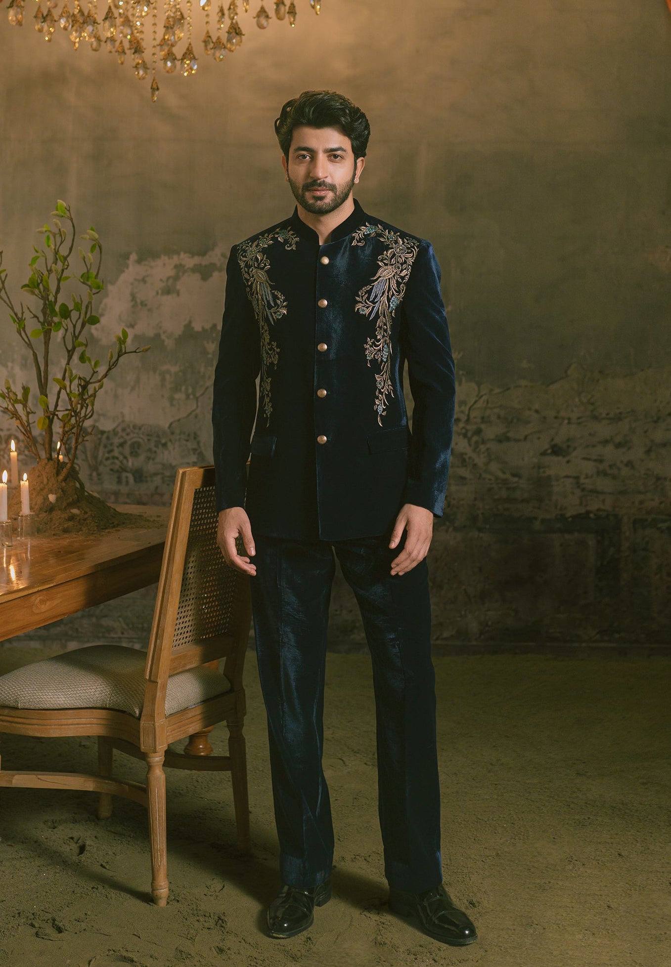 A man wearing a dark navy blue embroidered traditional Indian formal suit with gold buttons and silver mirror-work details on the chest, standing in a dimly lit room with a chandelier, table, and candles.