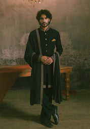 A man wearing a traditional black sherwani with ornate silver-gray border trim and gold buttons.