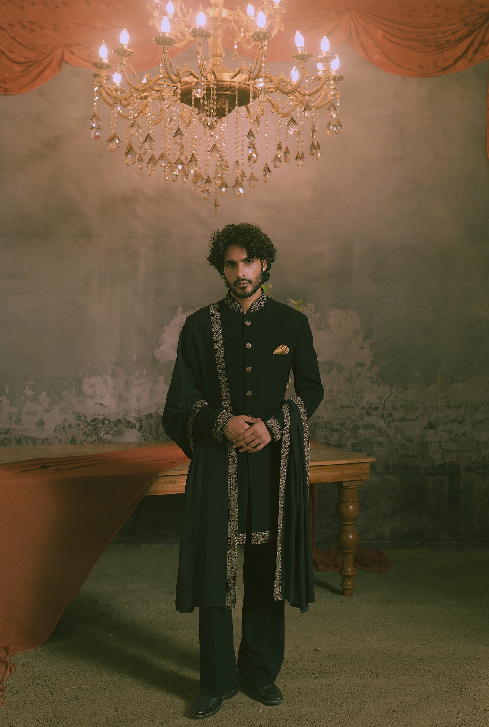 A man stands in a dimly lit room wearing a long black traditional outfit with ornate silver-gray trim, dark trousers, and a small gold pocket square.