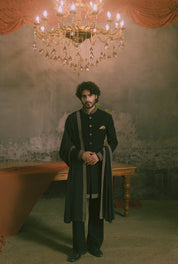 A man stands in a dimly lit room wearing a long black traditional outfit with ornate silver-gray trim, dark trousers, and a small gold pocket square.