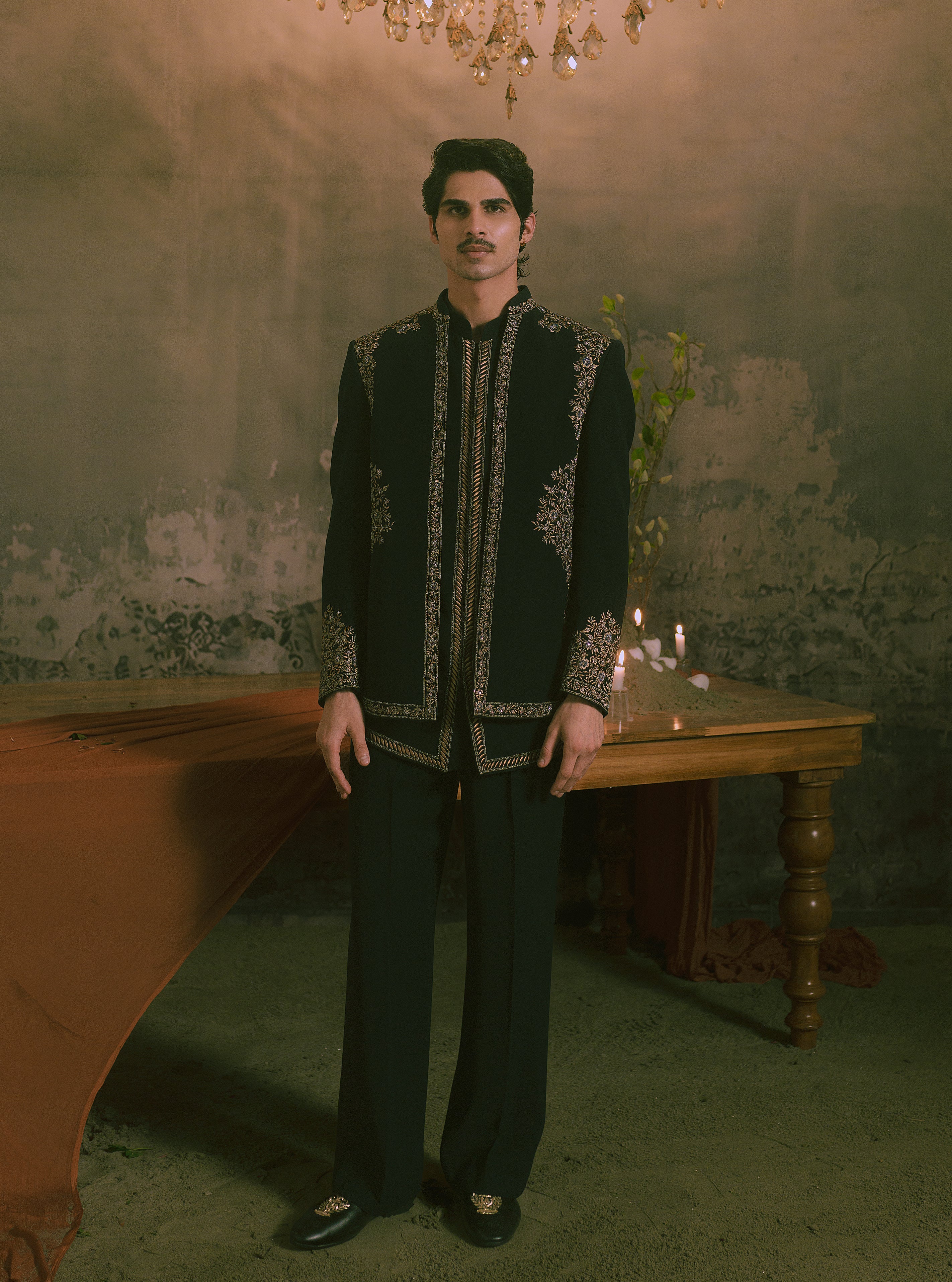 A man wearing a black traditional embroidered jacket and matching trousers stands in a dimly lit room with a rustic wall, a wooden table, and a chandelier overhead.