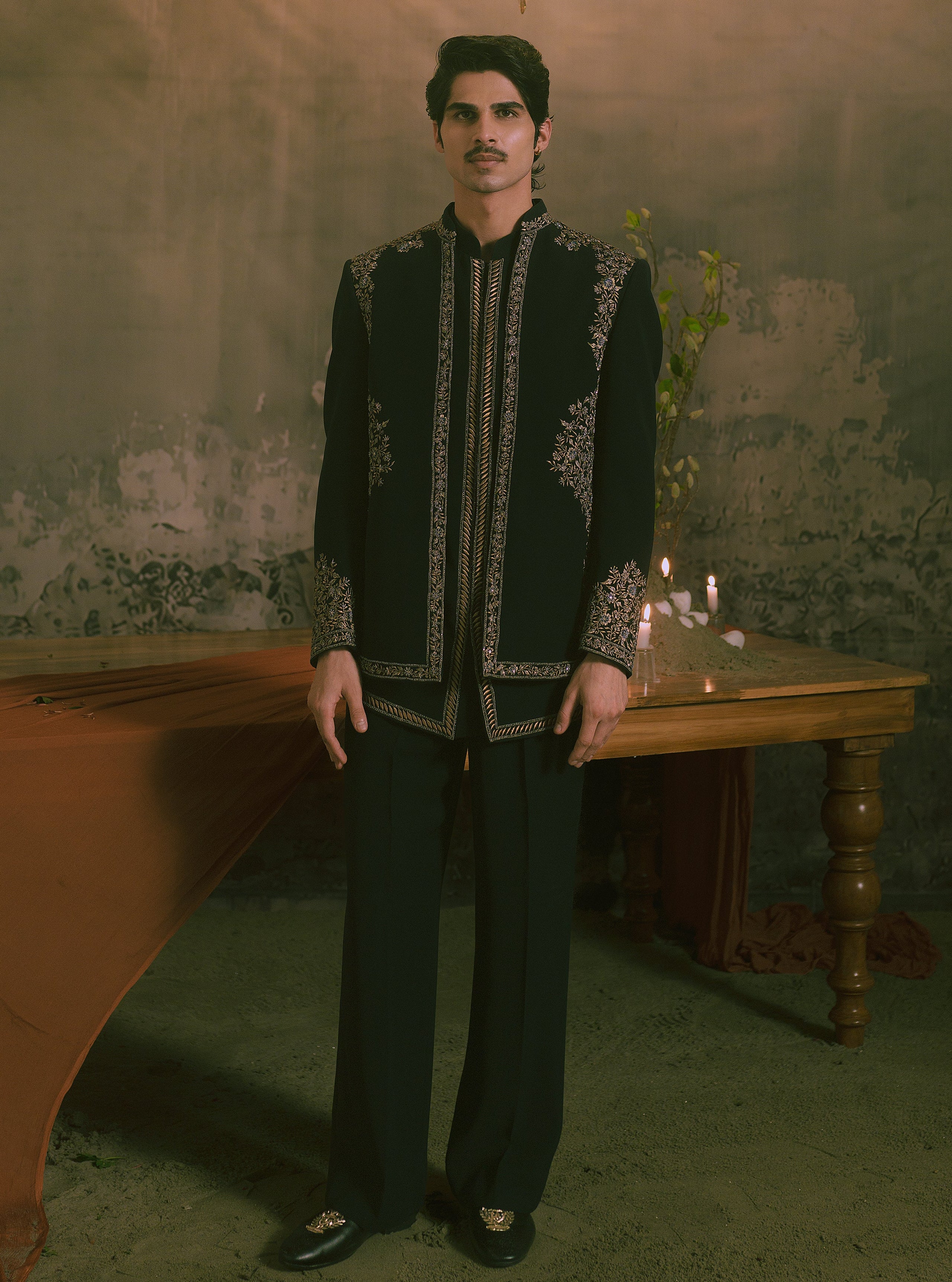A man wearing a black traditional embroidered jacket and matching trousers stands in a dimly lit room with a rustic wall, a wooden table, and a chandelier overhead.