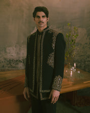 A man wearing a black traditional embroidered jacket with ornate gold and silver detailing, standing in a rustic room with candles and a textured wall.
