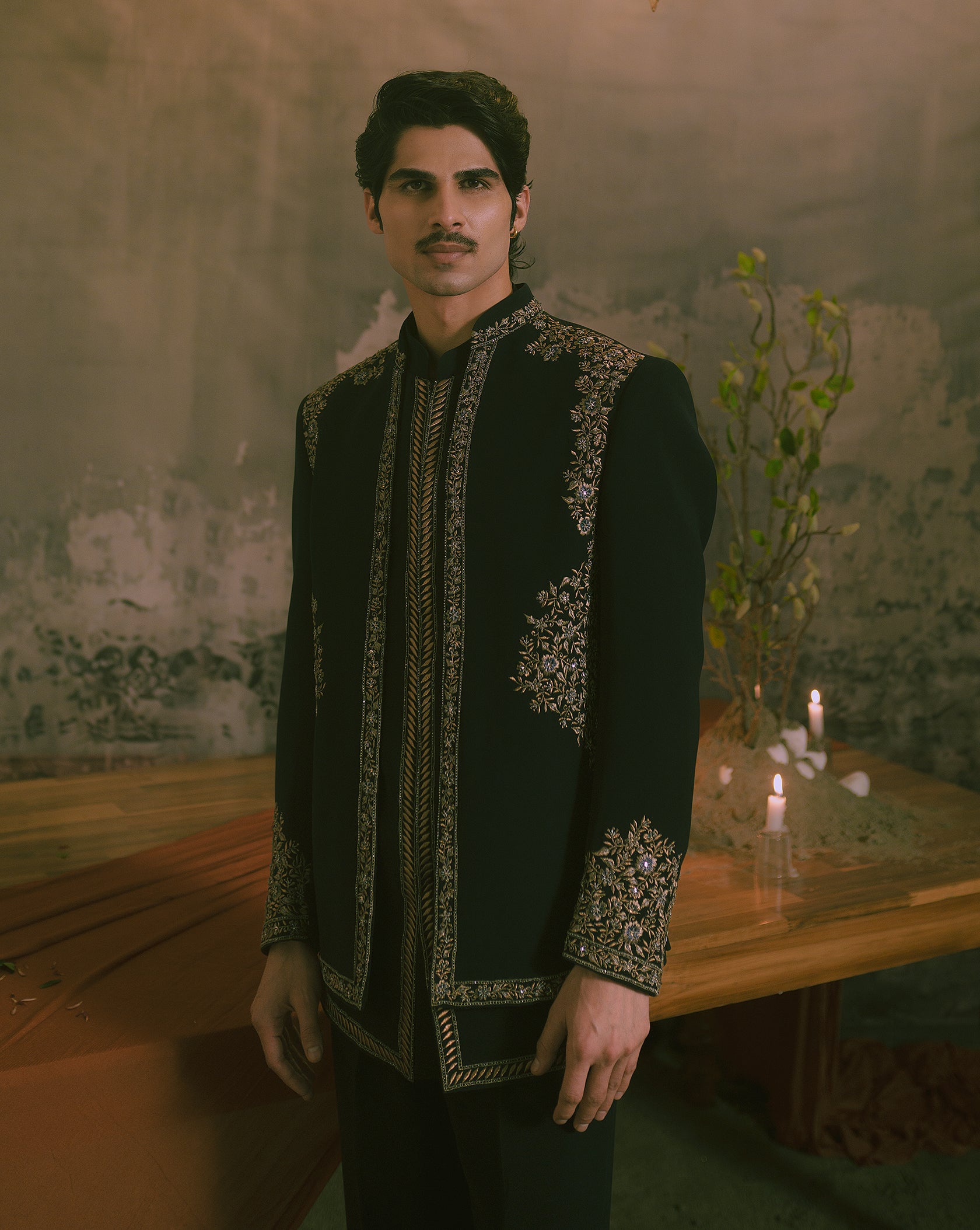 A man wearing a black traditional embroidered jacket with ornate gold and silver detailing, standing in a rustic room with candles and a textured wall.