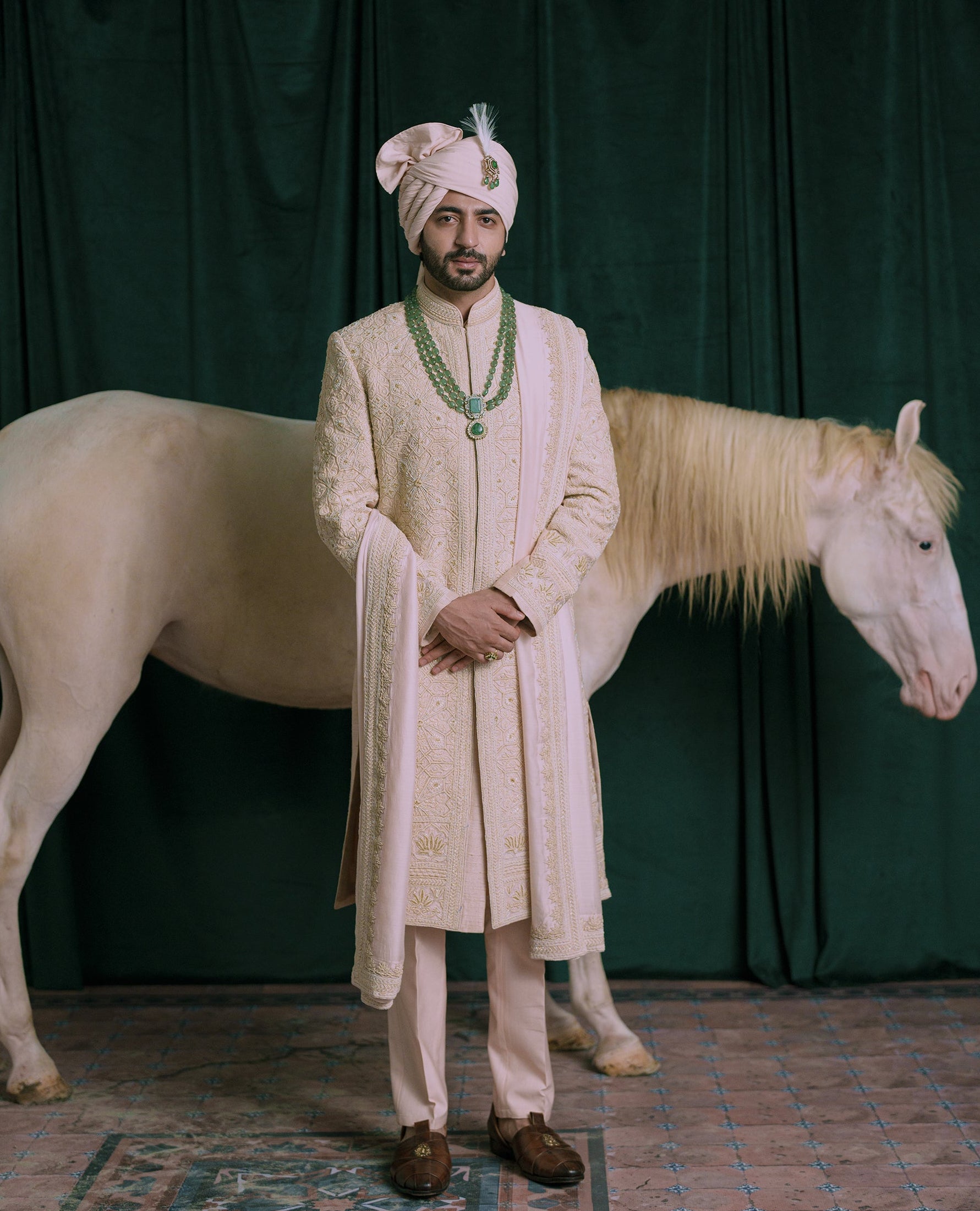 A man wearing a traditional pink hand embroidered sherwani and matching turban stands in front of a dark green draped backdrop with a white horse behind him.