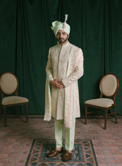 A man is wearing a traditional Indian outfit. He is dressed in a cream-colored embroidered sherwani with light green trousers & a stole to complete the look.