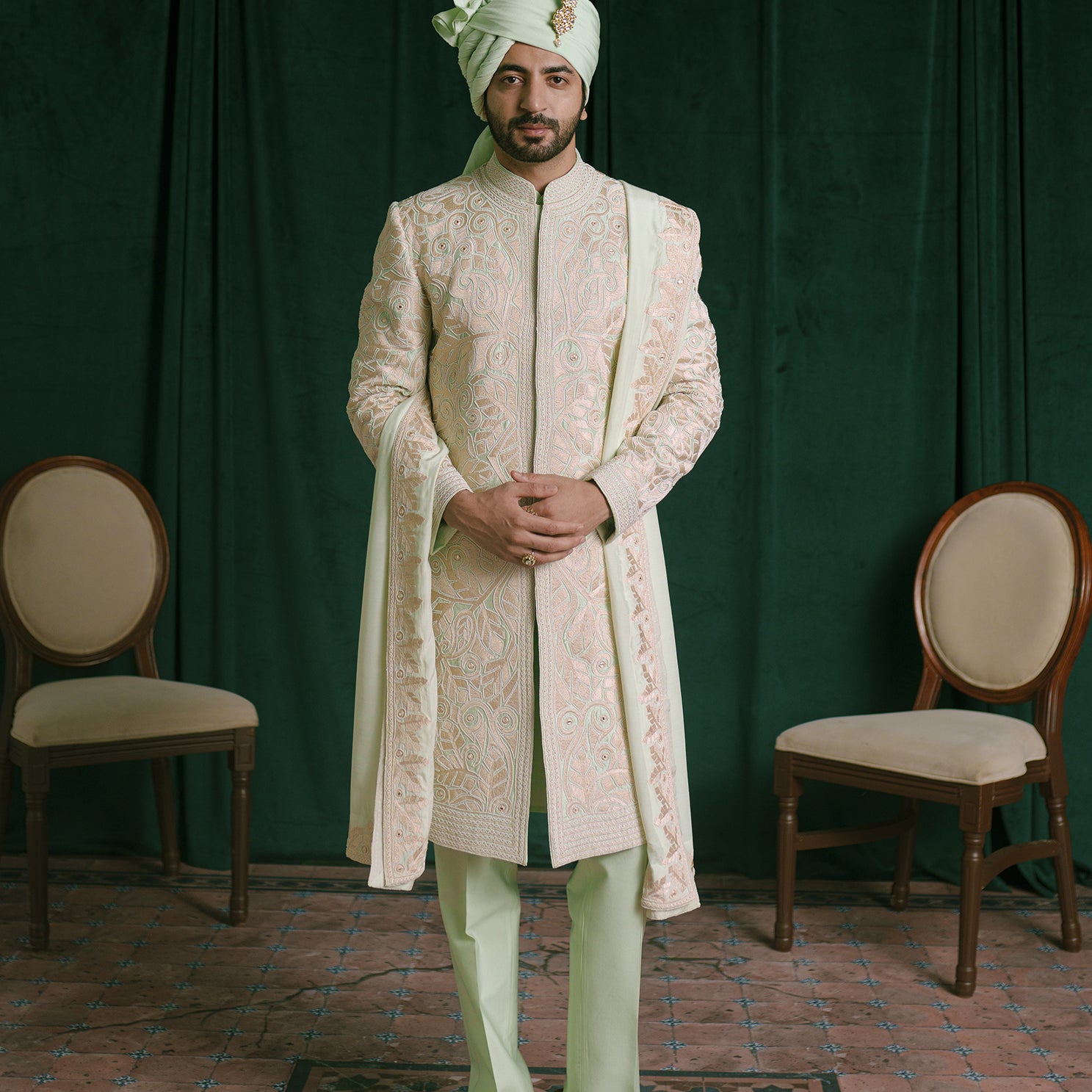 A man stands in a studio wearing a traditional Indian ethnic wear meant for wedding events, featuring a light green colored embroidered sherwani jacket, matching trousers, a light green turban, and a stole to complete the look.