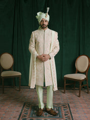A man stands in a studio wearing a traditional Indian ethnic wear meant for wedding events, featuring a light green colored embroidered sherwani jacket, matching trousers, a light green turban, and a stole to complete the look.
