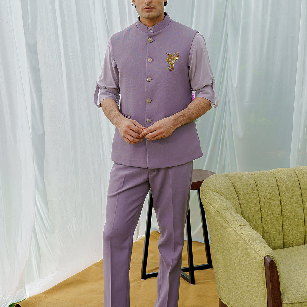 Man wearing a lavender traditional outfit standing in a room with a green chair and white curtains.