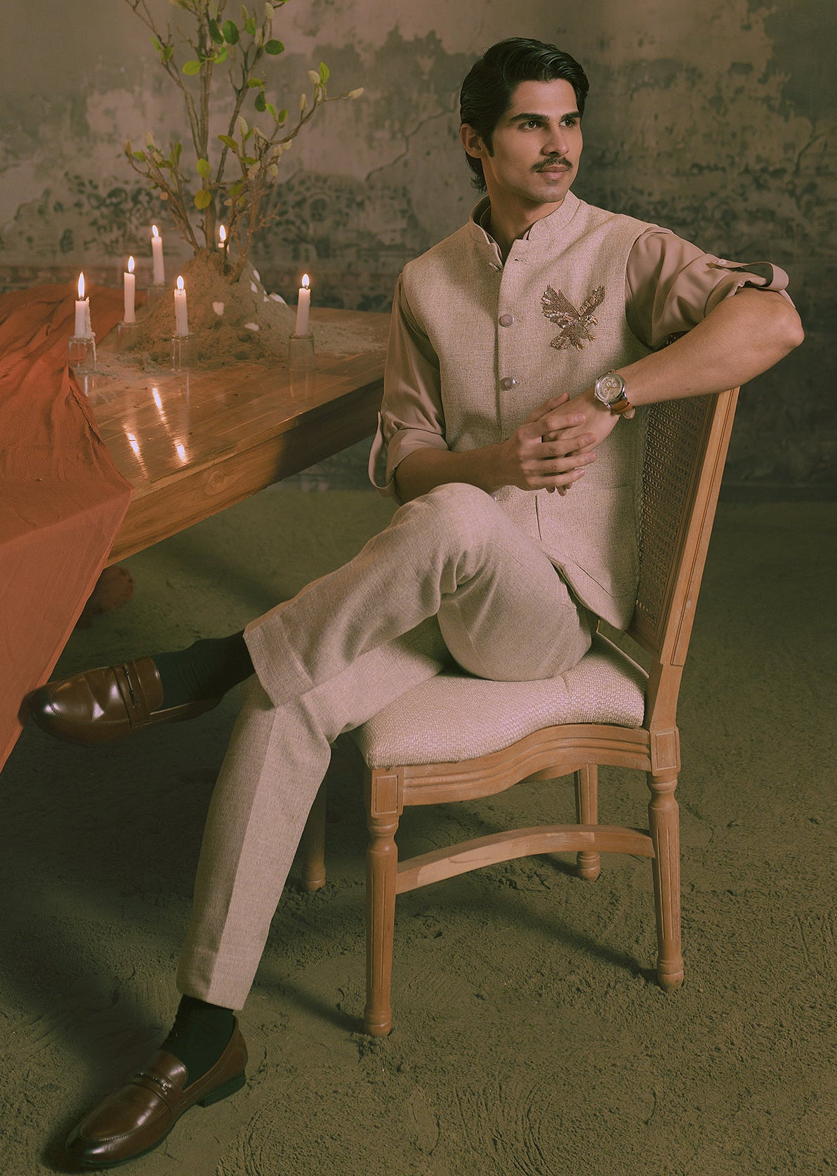 A man wearing a light beige traditional Indian outfit with an hand embroidered eagle sits on a wooden chair in a dimly lit, vintage-style room with a chandelier, table, and candles.