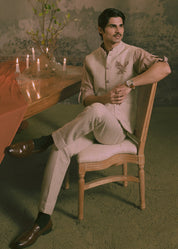 A man wearing a light beige traditional Indian outfit with an hand embroidered eagle sits on a wooden chair in a dimly lit, vintage-style room with a chandelier, table, and candles.