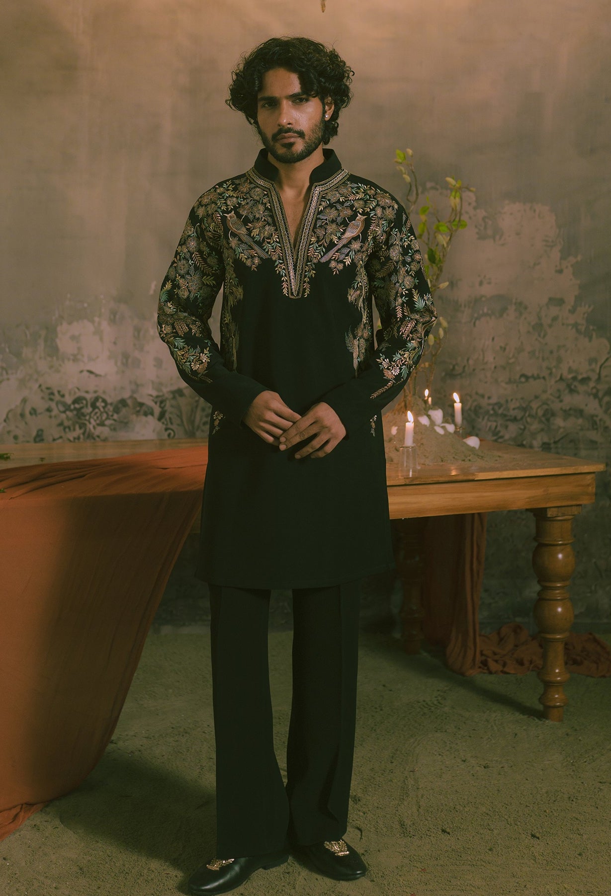 A man wearing a black traditional kurta with ornate gold and silver embroidery on the sleeves and collar, paired with black trousers.