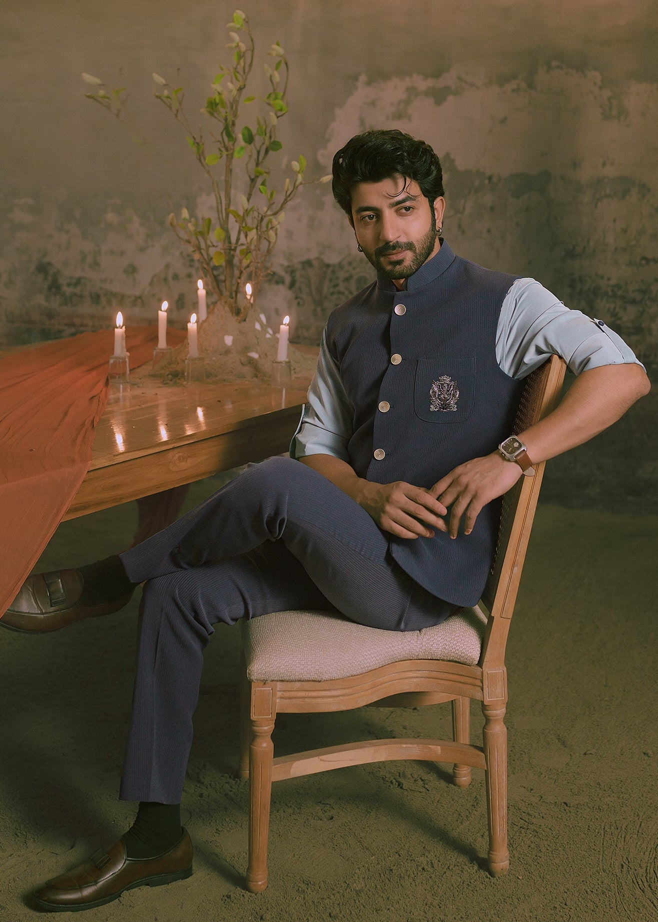A man wearing a navy blue traditional Indian nehru jacket outfit that has hand embroidery with a light blue shirt sits on a wooden chair in a rustic room decorated with a large chandelier, draped orange fabric, and candles.