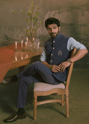 A man wearing a navy blue traditional Indian nehru jacket outfit that has hand embroidery with a light blue shirt sits on a wooden chair in a rustic room decorated with a large chandelier, draped orange fabric, and candles.